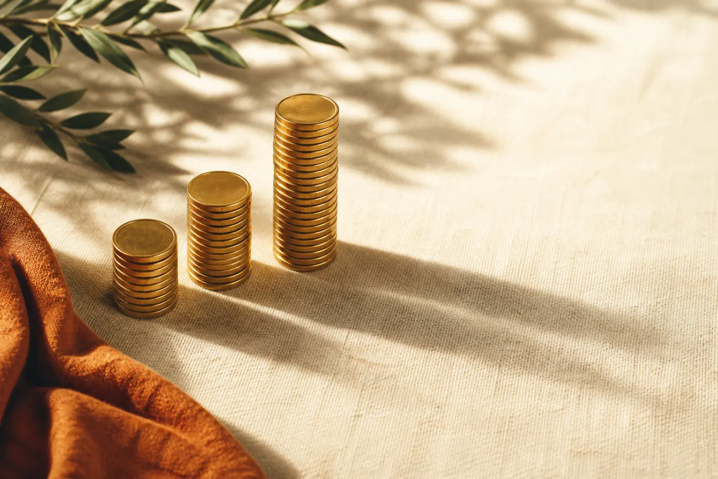 Three escalating stacks of polished gold coins on a cream linen surface, dappled with leaf shadows.